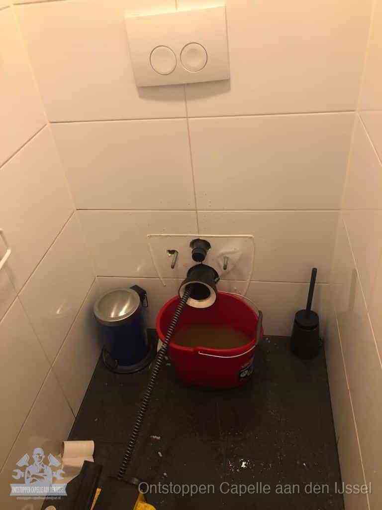 Een ontstopte wand-wc met verwijderde toiletpot waar een ontstoppingsveer in de wanduitgang verdwijnt, het achterhalen waar de verstopping zit in Capelle aan den IJssel