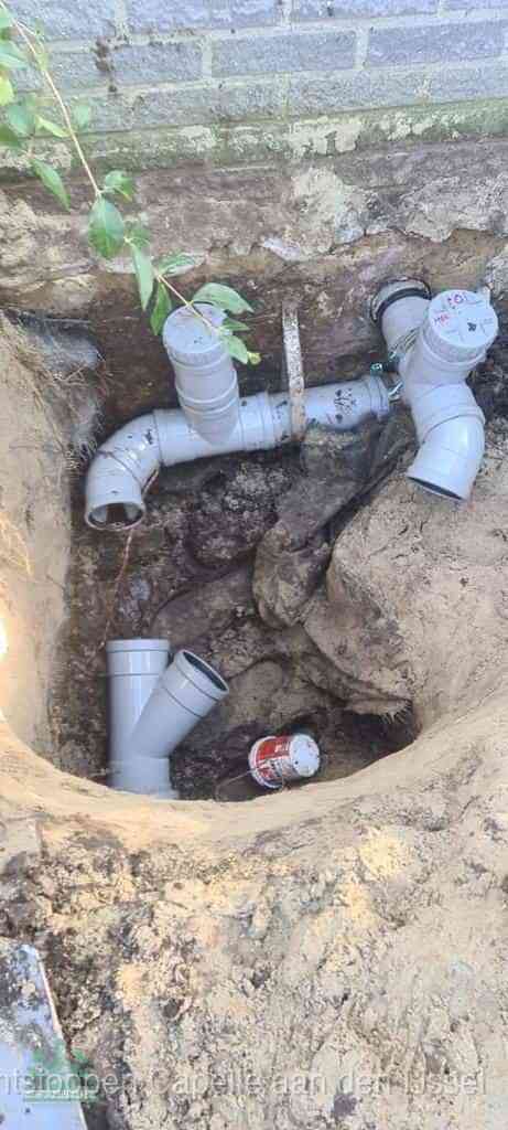 Een opgegraven bouwput langs een huismuur met grijze PVC rioolbuizen die op het hoofdriool worden aangesloten, typisch leidingwerk per wijk in Capelle aan den IJssel